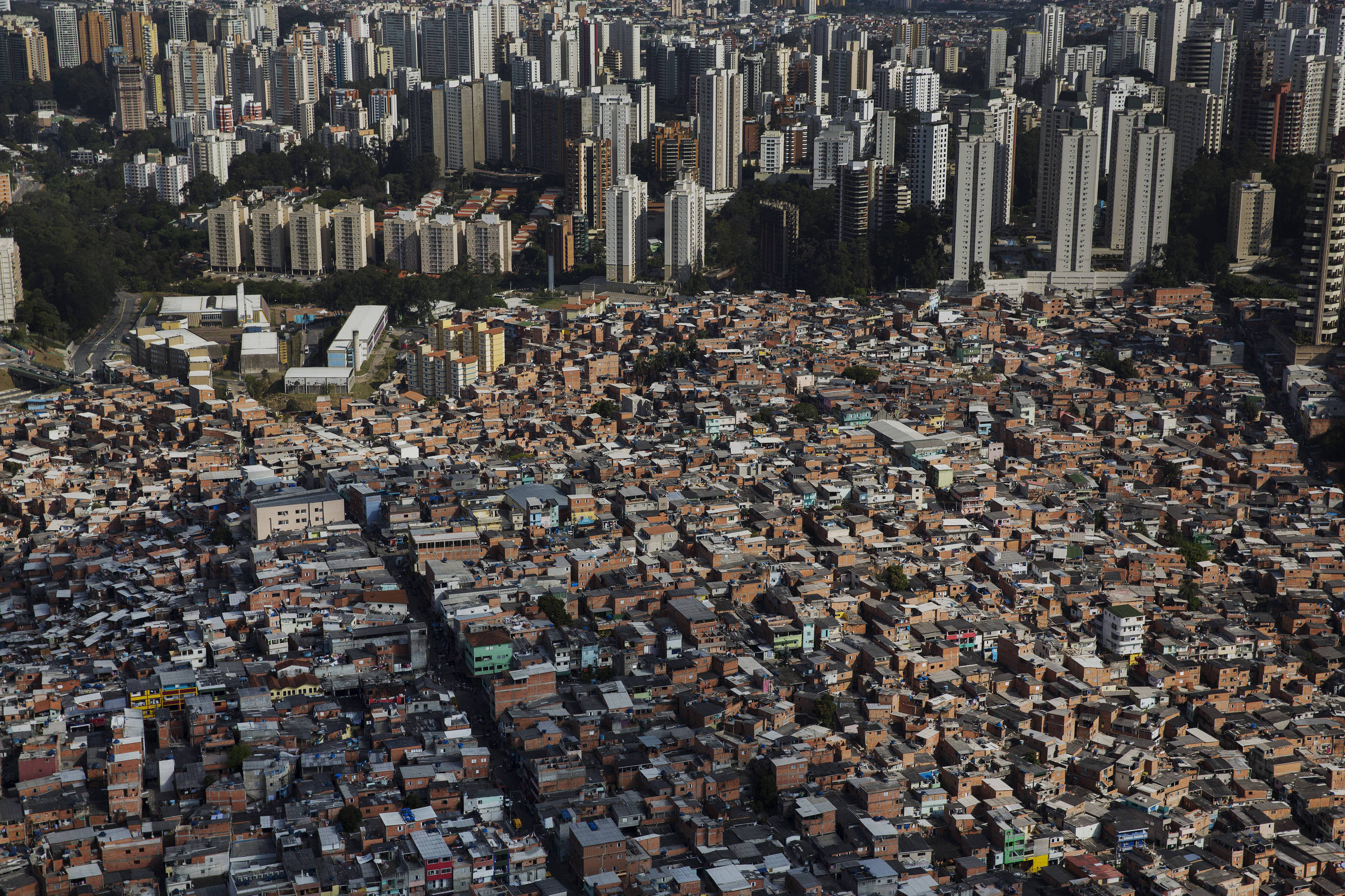 fotografia colorida mostra, vista de cima, a favela de Paraisópolis. Ao fundo, há os prédios de um bairro nobre, de forma que a fronteira entre a favela e o restante da região é nítida