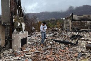 A imagem mostra um jovem em meio a escombros de uma construção destruída por um incêndio. O chão está coberto de destroços, incluindo pedaços de telhas e materiais de construção. Ao fundo, há uma estrutura parcialmente queimada e uma paisagem montanhosa com nuvens escuras. O céu está nublado, indicando um clima sombrio.