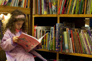 Uma menina com cabelo cacheado e uma faixa na cabeça está sentada em uma prateleira de livros, lendo um livro de capa rosa. Ela usa um casaco rosa claro e parece concentrada na leitura. Ao fundo, há várias prateleiras cheias de livros coloridos, com títulos visíveis como