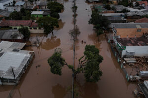 Imagem mostra uma avenida totalmente alagada. Aparece vegetação no canteiro central e casas ao redor, também com os quintais cheios d