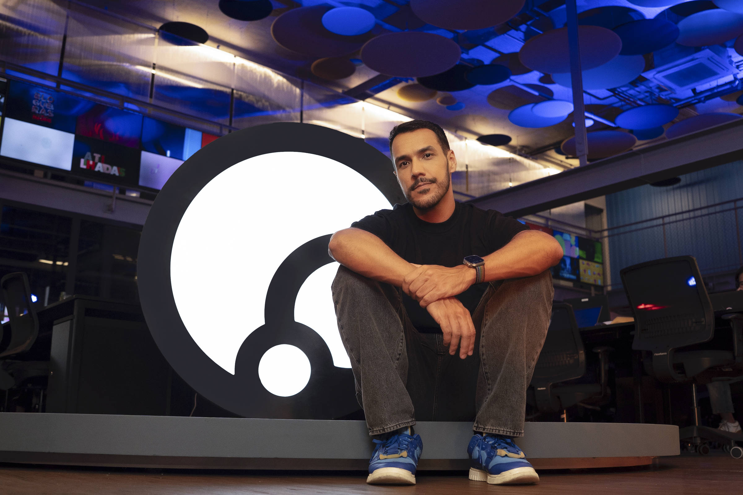 Um homem sentado em frente a um grande logotipo circular em um ambiente de escritório moderno. O homem está usando uma camiseta preta, calças escuras e tênis azuis. O fundo apresenta uma iluminação azul e várias telas visíveis com conteúdo variado.