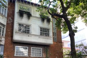 A imagem mostra uma casa de três andares, construída em tijolos, com uma fachada branca na parte superior. A casa possui janelas com grades e está cercada por um portão branco. Há vegetação ao redor, incluindo uma árvore à direita e plantas na base da casa. O ambiente é urbano, com calçada visível em primeiro plano.