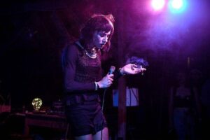 A imagem mostra um artista em uma apresentação ao vivo, segurando um microfone em uma mão e um cigarro na outra. O ambiente é escuro, iluminado por luzes coloridas, principalmente em tons de roxo e azul. O artista está vestido com uma blusa preta e uma saia curta, com cabelo escuro e liso. Ao fundo, é possível ver equipamentos de som e algumas pessoas assistindo à performance.