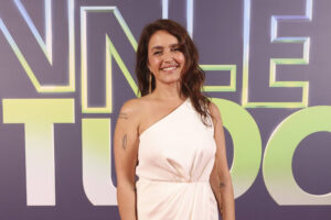 Uma mulher está posando em um evento de gala, vestindo um vestido longo branco com um ombro só. Ela sorri e está em pé sobre um tapete vermelho, com um fundo que apresenta o texto