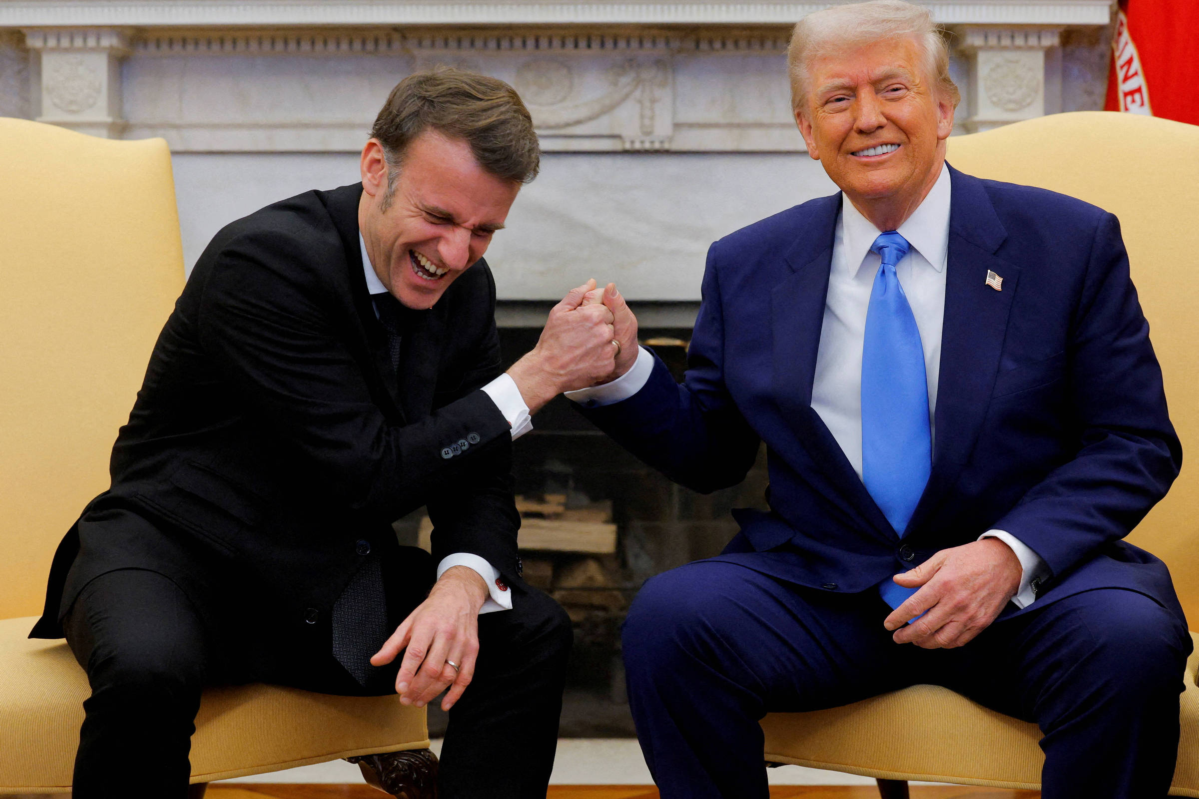 A imagem mostra dois homens sentados em cadeiras em um ambiente formal. Um deles está rindo e segurando a mão do outro em um gesto de cumprimento. O homem à esquerda está vestido com um terno escuro e uma camisa clara, enquanto o homem à direita usa um terno azul e uma gravata azul clara. Ao fundo, há uma lareira e uma decoração clássica.