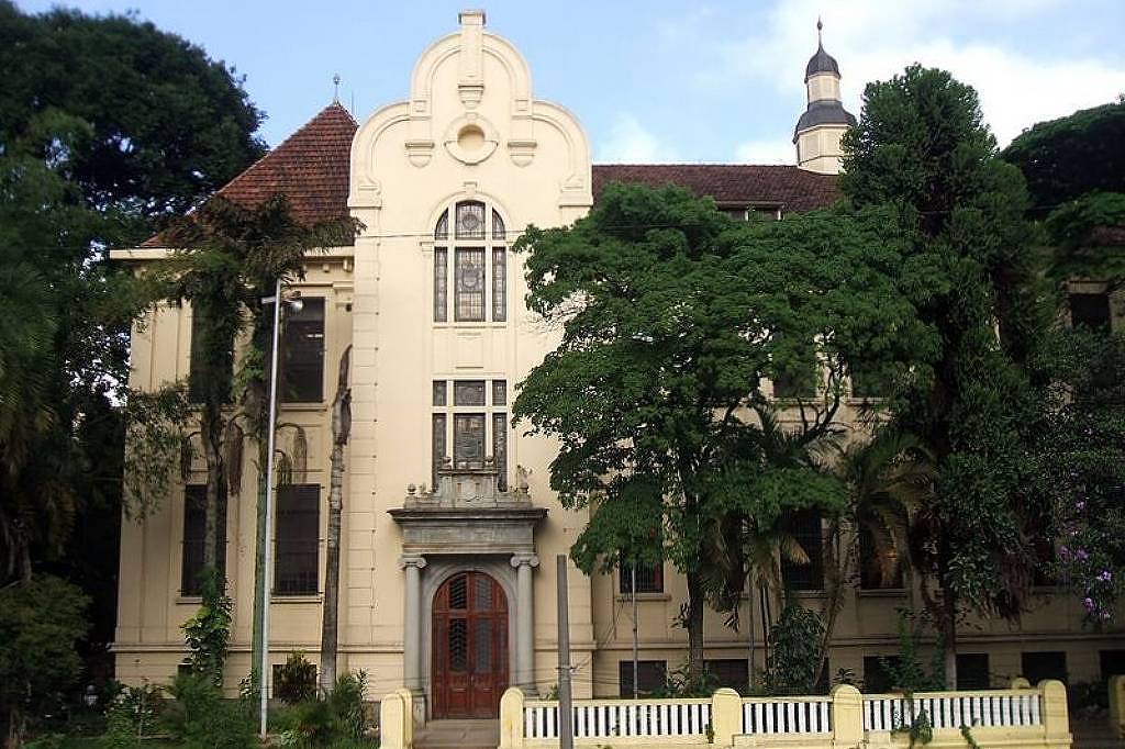 A imagem mostra um edifício histórico de arquitetura clássica, com paredes em tom claro e detalhes ornamentais. O prédio possui um telhado inclinado com uma torre no topo e janelas grandes. À frente, há um jardim com árvores e um muro baixo de cor clara. O céu está parcialmente nublado.