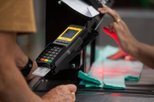 A imagem mostra um terminal de pagamento em um caixa, com uma mão segurando um cartão e outra mão interagindo com o terminal. O terminal é preto com um visor amarelo e botões coloridos. Ao fundo, há um balcão e alguns itens de compras visíveis.