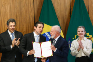A imagem mostra uma cerimônia de assinatura de um documento oficial. Quatro pessoas estão presentes: um homem à esquerda, com cabelo grisalho e terno escuro, aplaudindo; um homem no centro, com cabelo escuro e terno claro, segurando um documento; um homem à direita, com cabelo grisalho e terno escuro, sorrindo e segurando o documento; e uma mulher à direita, com cabelo liso e claro, vestindo uma blusa clara, também aplaudindo. Ao fundo, há bandeiras do Brasil.