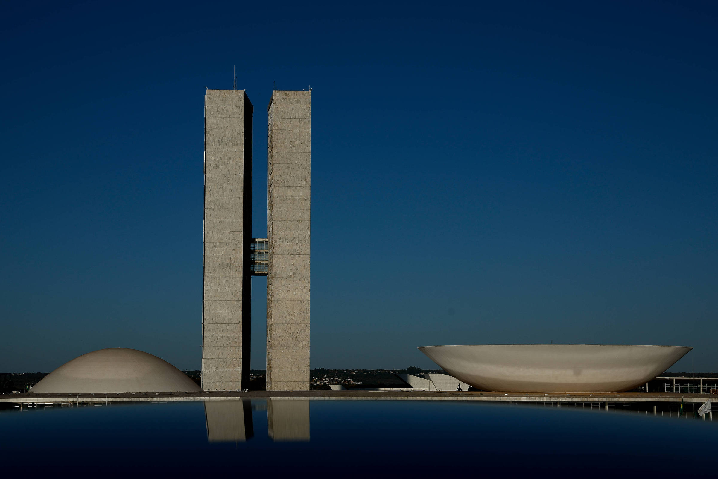 A imagem mostra o Congresso Nacional do Brasil, com suas duas torres retangulares e as cúpulas em forma de meia-lua e de prato. O céu está limpo e azul, refletindo na superfície da água em primeiro plano.