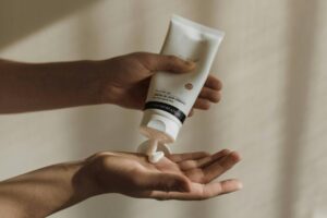 Uma mão está segurando um tubo de creme hidratante, enquanto a outra mão recebe uma quantidade do produto. O creme é de cor branca e está sendo dispensado do tubo. O fundo é neutro e suave, sugerindo um ambiente calmo.