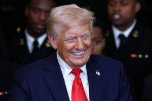 A imagem mostra um homem sorridente, vestido com um terno escuro e uma gravata vermelha, segurando uma bola de futebol americano. Ao seu lado, um jovem oficial naval, vestido com um uniforme escuro, também sorri. Ao fundo, é possível ver outros homens em uniformes navais, todos aparentando estar em um ambiente formal. A atmosfera é de celebração e camaradagem.