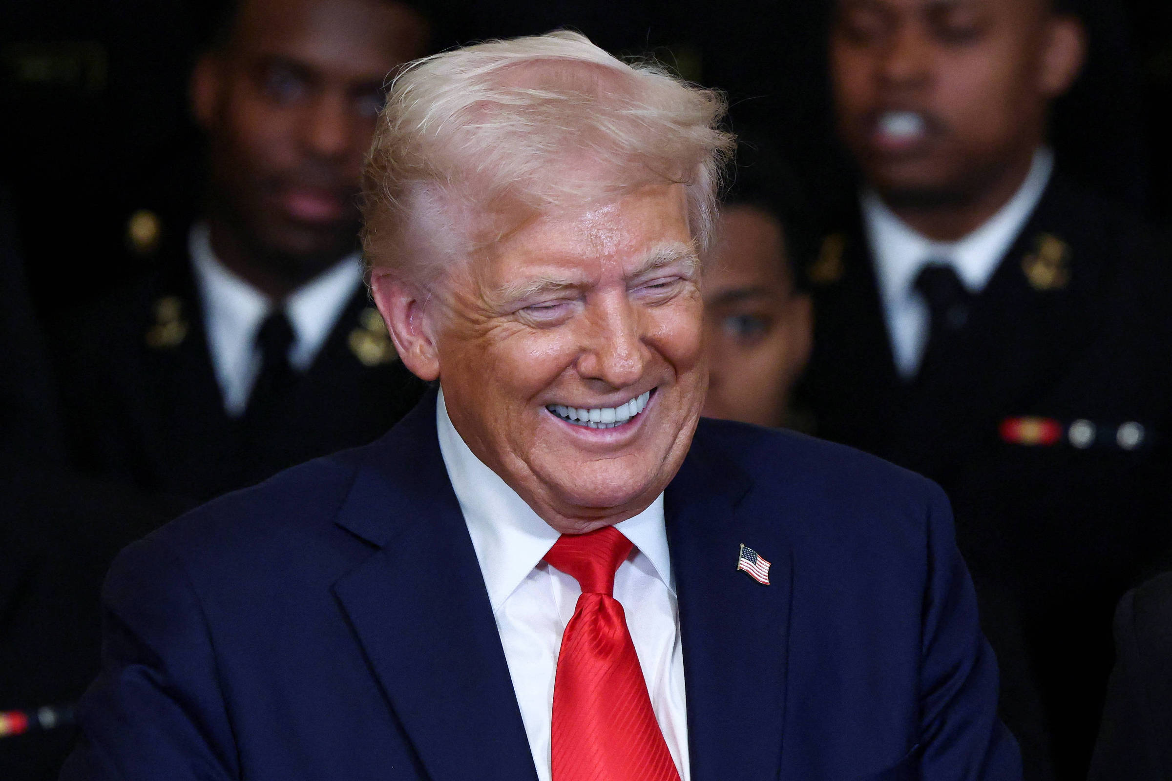 A imagem mostra um homem sorridente, vestido com um terno escuro e uma gravata vermelha, segurando uma bola de futebol americano. Ao seu lado, um jovem oficial naval, vestido com um uniforme escuro, também sorri. Ao fundo, é possível ver outros homens em uniformes navais, todos aparentando estar em um ambiente formal. A atmosfera é de celebração e camaradagem.