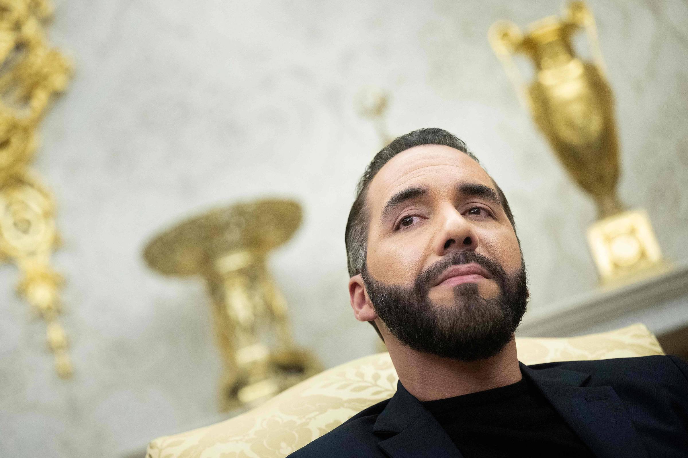 Um homem com barba e cabelo bem arrumado está sentado em um sofá, olhando para o lado. Ele usa uma camisa preta e um blazer escuro. Ao fundo, há elementos decorativos dourados, incluindo vasos e ornamentos, que sugerem um ambiente luxuoso. A iluminação é suave, destacando o rosto do homem.