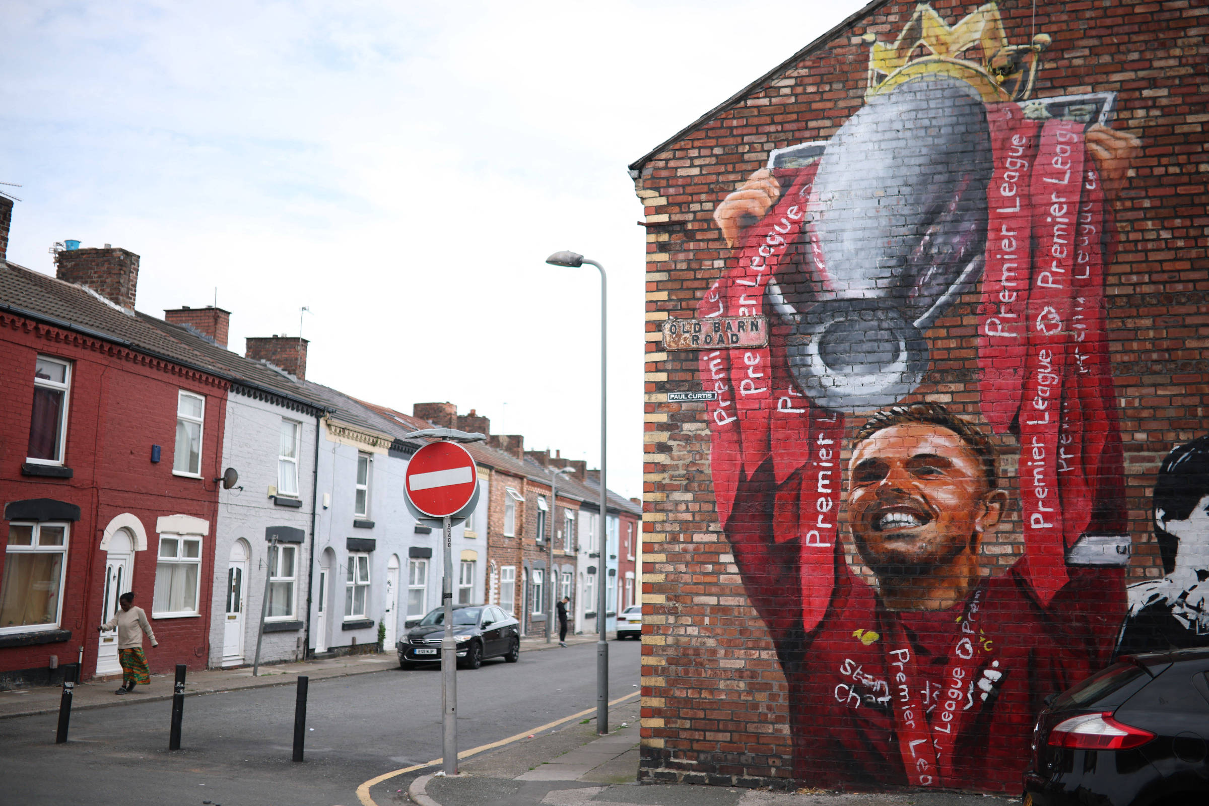 A imagem mostra um mural em uma parede de tijolos, representando um jogador de futebol segurando um troféu. O mural é colorido, com predominância de vermelho e detalhes em branco. Ao fundo, há uma rua com casas de um lado e um sinal de trânsito indicando proibição de entrada. O ambiente é urbano e parece ser uma área residencial.