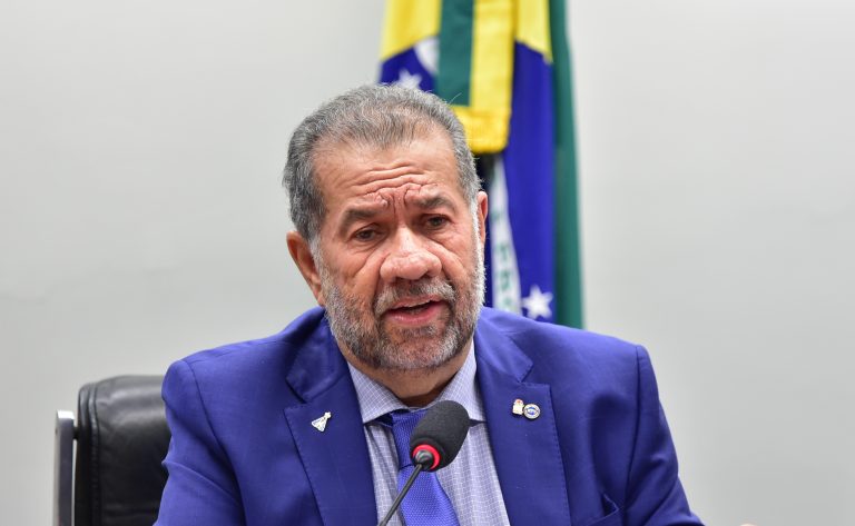 Um homem branco com barba e cabelo grisalho usa terno e fala ao microfone