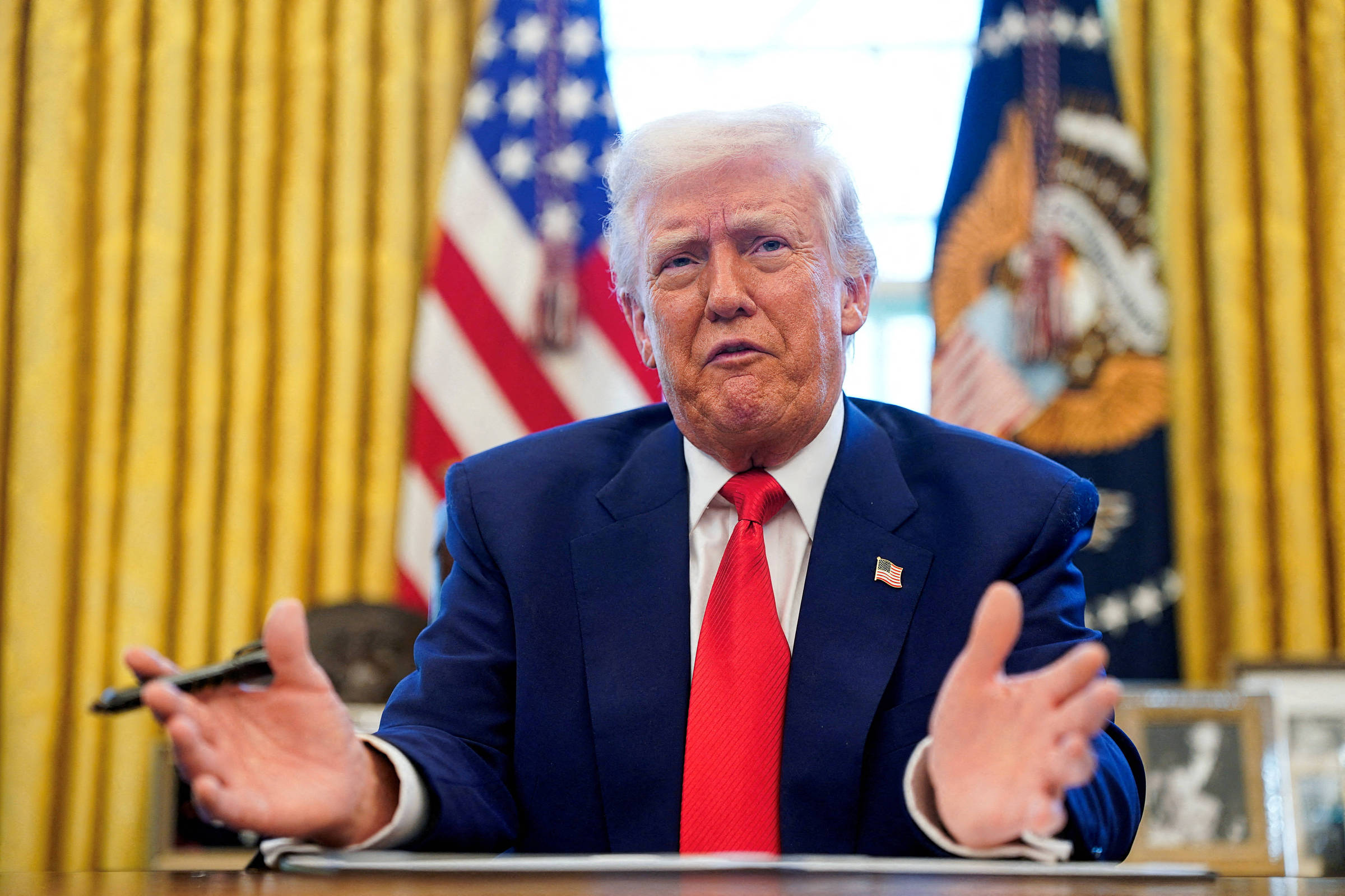 Um homem com cabelo loiro e terno escuro está sentado em uma mesa no Salão Oval. Ele está gesticulando com as mãos e parece estar falando. Ao fundo, há cortinas douradas e bandeiras dos Estados Unidos. Uma mesa com objetos decorativos está visível à sua frente.