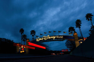 A imagem mostra o arco de entrada do Walt Disney World, iluminado à noite. O céu está nublado e escuro, com algumas palmeiras visíveis ao lado do arco. O nome