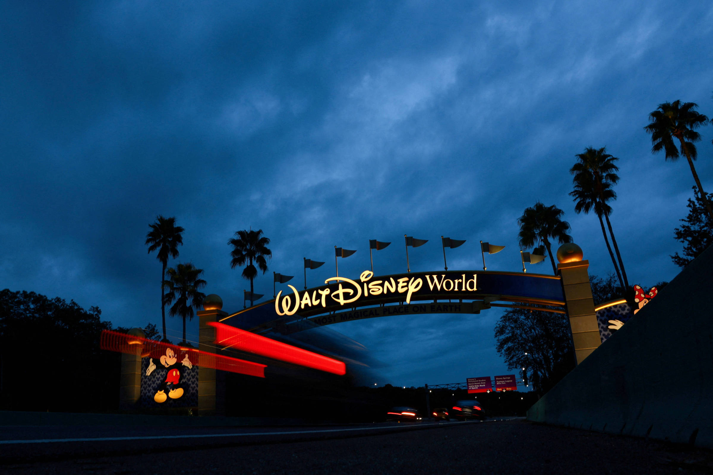 A imagem mostra o arco de entrada do Walt Disney World, iluminado à noite. O céu está nublado e escuro, com algumas palmeiras visíveis ao lado do arco. O nome