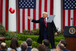A imagem mostra um homem de terno escuro e gravata vermelha, em um evento ao ar livre, lançando um boné vermelho. Ao fundo, há bandeiras dos Estados Unidos e um público assistindo. O homem está em frente a um púlpito com o selo presidencial. A cena é iluminada e parece ser um momento de destaque durante o evento.