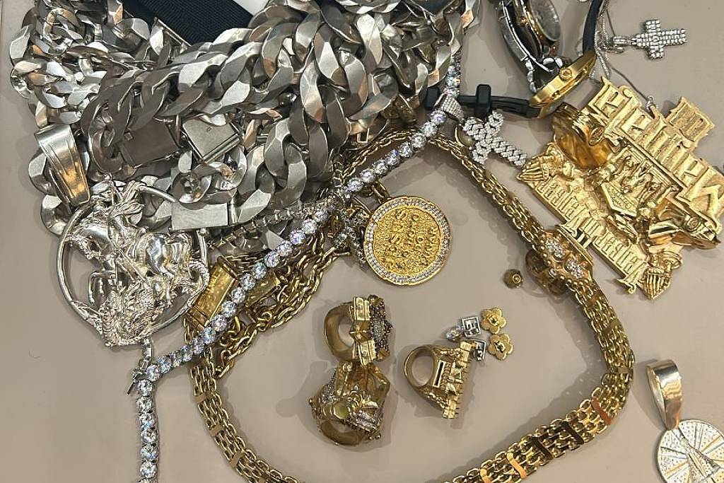 A imagem mostra uma variedade de joias e acessórios dispostos sobre uma mesa. Há correntes de prata e ouro, pingentes, um relógio dourado, um chaveiro de carro e uma imagem de um carro Lamborghini. As joias incluem cruzes, medalhas e outros enfeites, com detalhes brilhantes e elaborados. O fundo é de madeira clara.