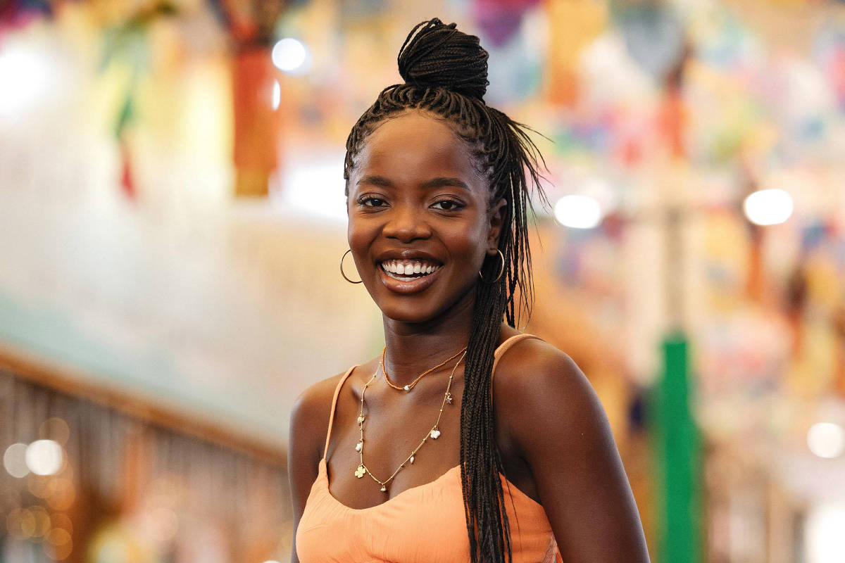A imagem mostra uma mulher sorridente em um ambiente interno decorado com bandeirinhas coloridas. Ela está usando um macacão laranja e segurando uma bolsa de palha. O fundo apresenta uma loja com produtos variados e uma iluminação suave.