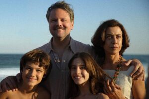 Uma família posando na praia, com o mar ao fundo. O pai, com um sorriso, está no centro, ao lado da mãe, que tem uma expressão séria. À esquerda, um menino sorridente e à direita, uma menina também sorridente. Todos estão vestidos de forma casual, com a luz do sol iluminando a cena.