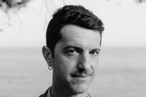 A imagem mostra um homem em um retrato em preto e branco. Ele está posicionado em frente a um fundo que parece ser o mar, com uma árvore ou folhagem visível na parte superior. O homem tem cabelo escuro e curto, um bigode e usa um brinco. Ele está vestido com uma camisa clara e um casaco. A expressão dele é séria e direta, olhando para a câmera.