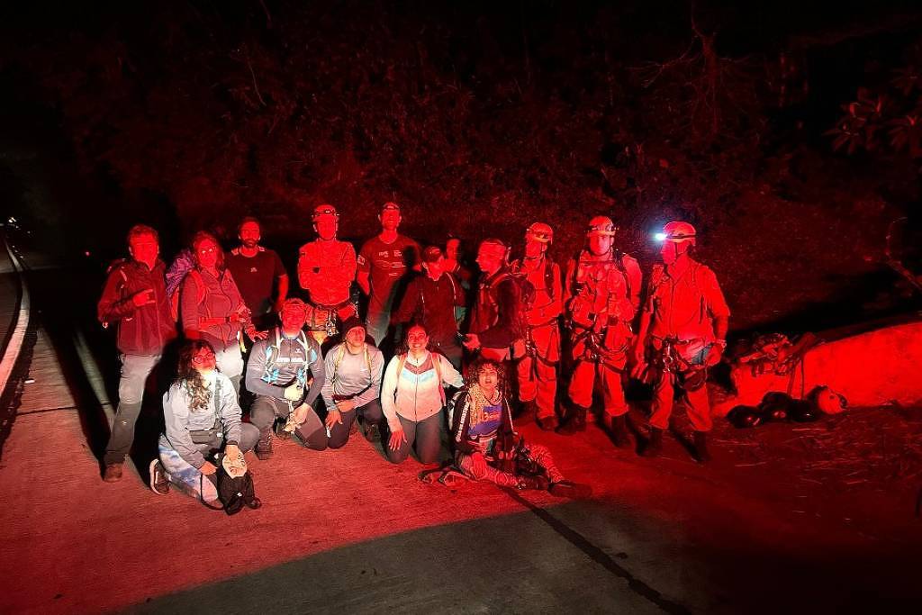 A imagem mostra um grupo de pessoas reunidas em uma estrada à noite, iluminadas por luzes vermelhas. O grupo é composto por homens e mulheres, alguns vestindo equipamentos de resgate, como cintos e capacetes. Eles estão posicionados em duas fileiras, com algumas pessoas agachadas na frente e outras em pé atrás. O ambiente ao redor é escuro, com vegetação visível ao fundo.