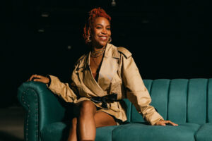 Uma mulher sorridente está sentada em um sofá azul. Ela usa um casaco bege e tem cabelo laranja em dreadlocks. A iluminação é suave e o fundo é escuro, destacando a modelo e o sofá.