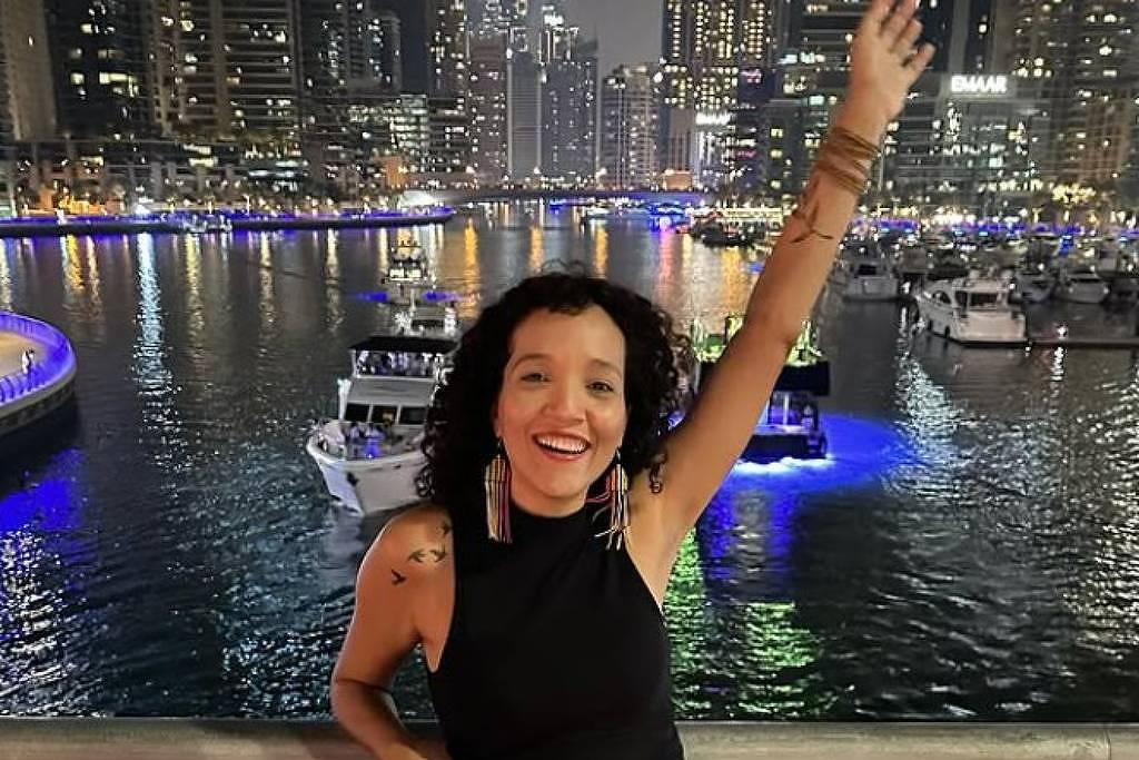 Uma mulher com cabelo cacheado e vestido preto está sorrindo e levantando o braço em um cenário noturno. Ao fundo, há um canal com barcos iluminados e arranha-céus de Dubai, refletindo luzes na água. A atmosfera é vibrante e festiva, com luzes azuis e brancas ao redor.