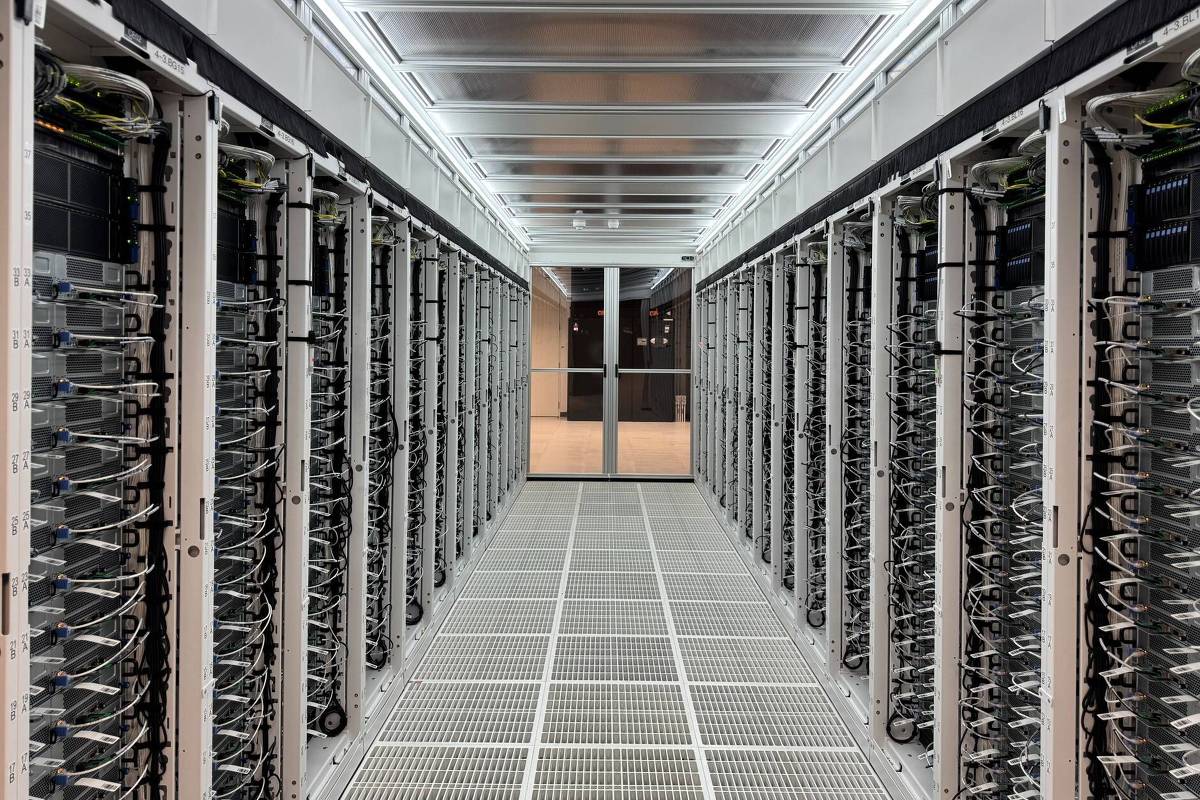 A imagem mostra um corredor de uma sala de servidores, com fileiras de servidores organizados em racks em ambos os lados. O ambiente é bem iluminado, com luzes no teto e um piso de grade. No fundo, há uma porta de vidro que leva a outra área.