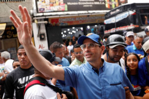 A imagem mostra um grupo de pessoas em uma rua movimentada. No centro, um homem com óculos e um boné azul acena com a mão. Ele está vestido com uma camisa azul e parece estar interagindo com a multidão ao seu redor. Ao fundo, há várias pessoas, algumas usando camisetas de marcas conhecidas, e uma loja com letreiros visíveis. O ambiente é urbano e parece haver um clima de agitação.
