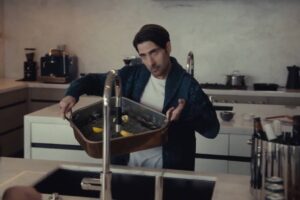 Um homem está em uma cozinha moderna, segurando uma bandeja com limões. Ele está vestido com uma camiseta branca e um casaco escuro. A cozinha possui armários brancos e uma bancada escura, com utensílios e eletrodomésticos visíveis ao fundo. O homem parece estar interagindo com outra pessoa fora do quadro.