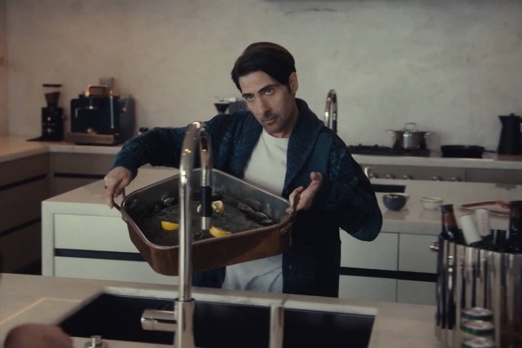 Um homem está em uma cozinha moderna, segurando uma bandeja com limões. Ele está vestido com uma camiseta branca e um casaco escuro. A cozinha possui armários brancos e uma bancada escura, com utensílios e eletrodomésticos visíveis ao fundo. O homem parece estar interagindo com outra pessoa fora do quadro.