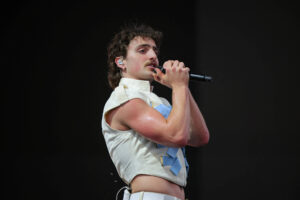 Um cantor está se apresentando em um palco. Ele usa uma camisa sem mangas de cor clara e calças brancas. O cantor tem cabelo cacheado e um bigode, segurando um microfone com as duas mãos enquanto canta. O fundo é escuro, sugerindo que ele está em um show ao vivo.