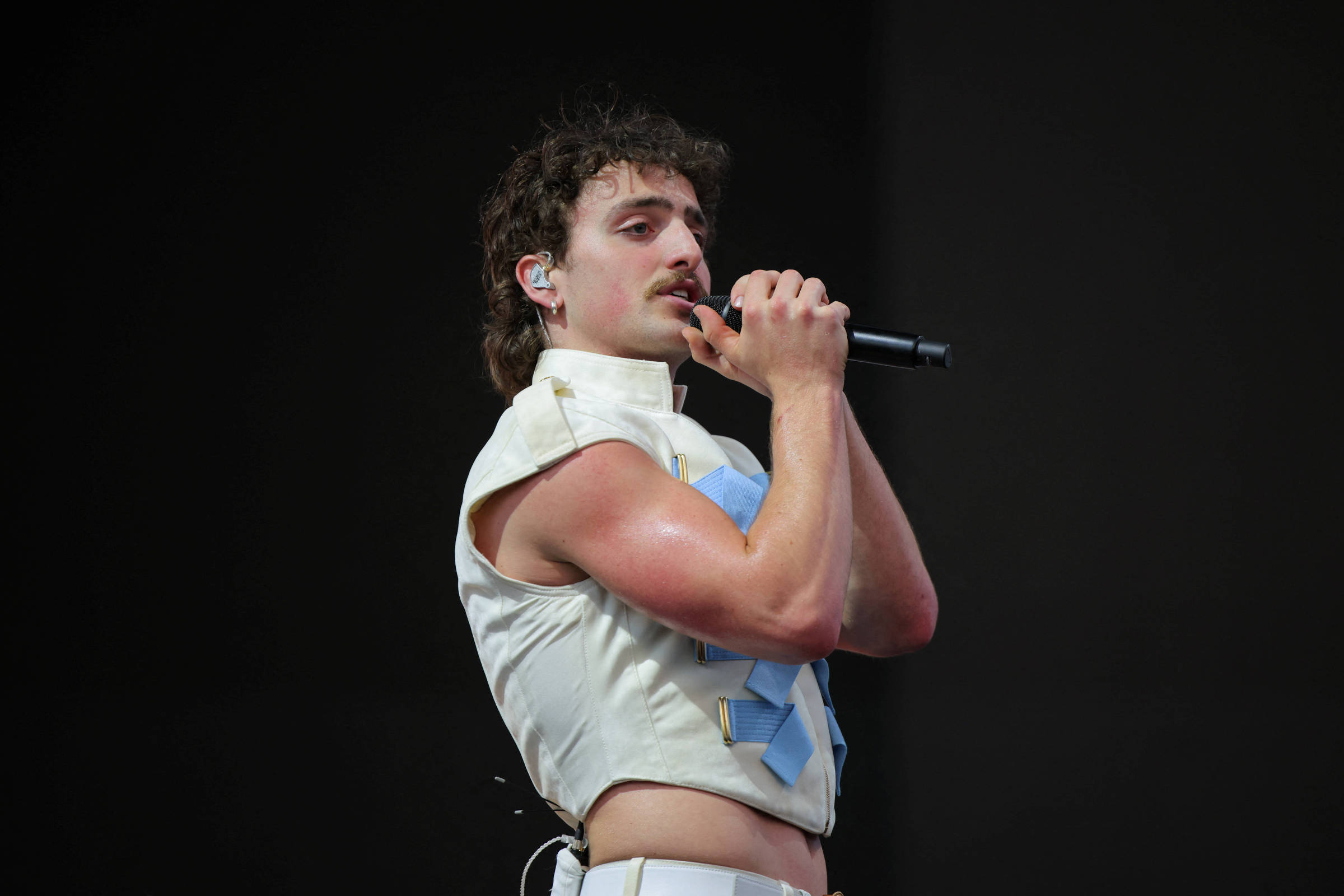 Um cantor está se apresentando em um palco. Ele usa uma camisa sem mangas de cor clara e calças brancas. O cantor tem cabelo cacheado e um bigode, segurando um microfone com as duas mãos enquanto canta. O fundo é escuro, sugerindo que ele está em um show ao vivo.