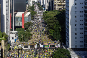 A imagem mostra uma grande multidão reunida em uma avenida de São Paulo, com pessoas vestindo camisetas amarelas. A cena é vista de cima, revelando uma longa fila de manifestantes que se estende pela rua, cercada por prédios altos e árvores. O clima parece ser de celebração ou protesto, com bandeiras e cartazes visíveis entre a multidão.