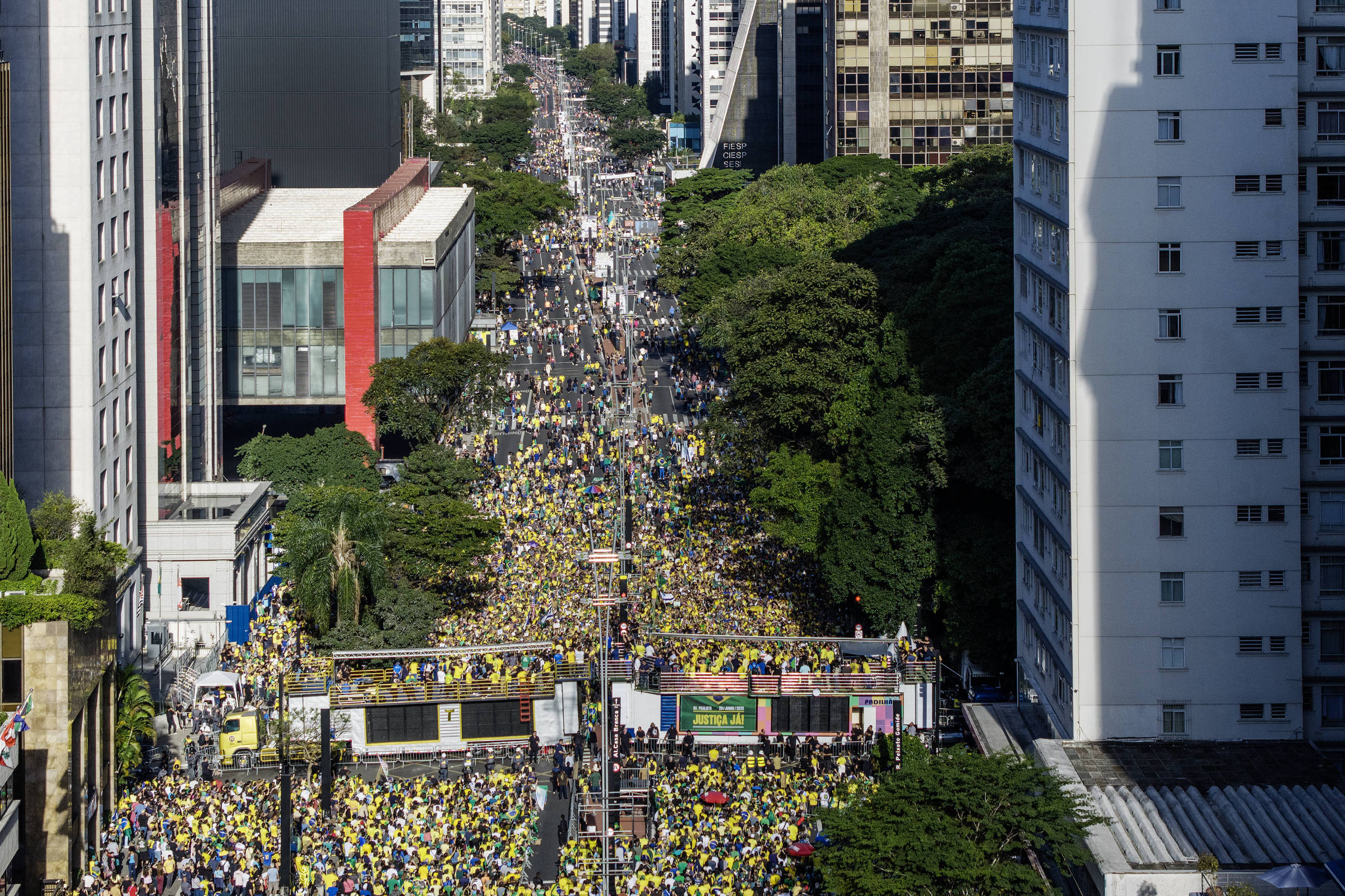 A imagem mostra uma grande multidão reunida em uma avenida de São Paulo, com pessoas vestindo camisetas amarelas. A cena é vista de cima, revelando uma longa fila de manifestantes que se estende pela rua, cercada por prédios altos e árvores. O clima parece ser de celebração ou protesto, com bandeiras e cartazes visíveis entre a multidão.