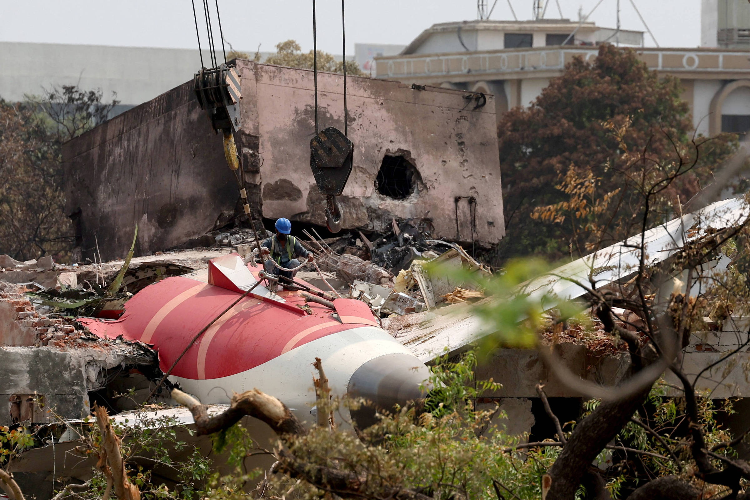 A imagem mostra os destroços de um avião em um local de demolição. Parte da fuselagem do avião, que é vermelha e branca, está visível entre os escombros de um edifício destruído. Um trabalhador, usando um capacete azul, está em cima dos destroços, enquanto guindastes estão levantando uma parte do edifício ao fundo, que apresenta buracos e danos significativos. A vegetação ao redor é visível, com árvores e arbustos.