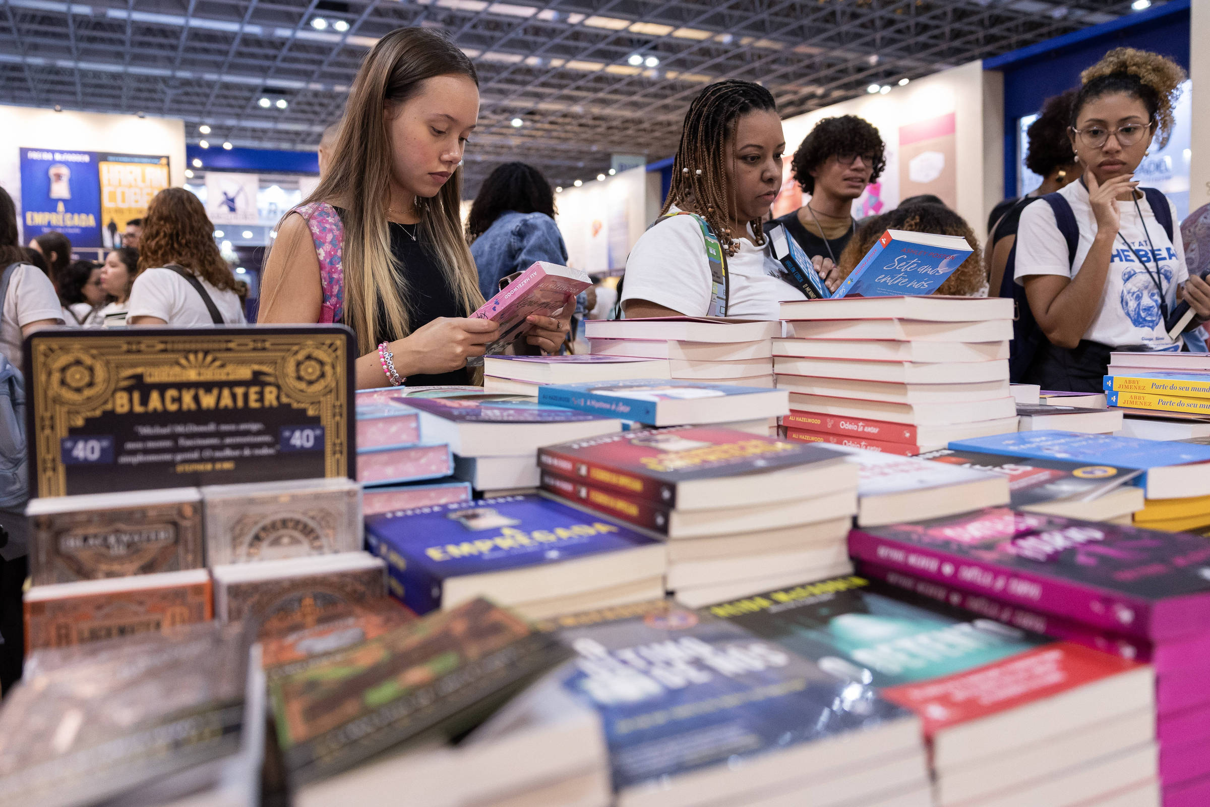 A imagem mostra um ambiente de feira do livro, com várias pessoas em pé ao redor de mesas repletas de livros. Algumas pessoas estão olhando para seus celulares, enquanto outras observam os livros expostos. Os livros têm capas coloridas e variadas, e há uma placa visível com a palavra