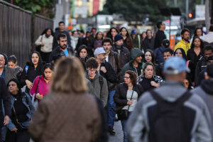 A imagem mostra uma grande multidão de pessoas caminhando em uma rua. As pessoas estão vestidas de maneira variada, algumas usando casacos e capuzes, indicando um clima mais frio. O cenário é urbano, com edifícios ao fundo e sinais de trânsito visíveis. A maioria das pessoas parece estar concentrada em seu caminho, algumas olhando para frente e outras para baixo. A iluminação sugere que a foto foi tirada durante o dia, possivelmente no início da manhã ou no final da tarde.