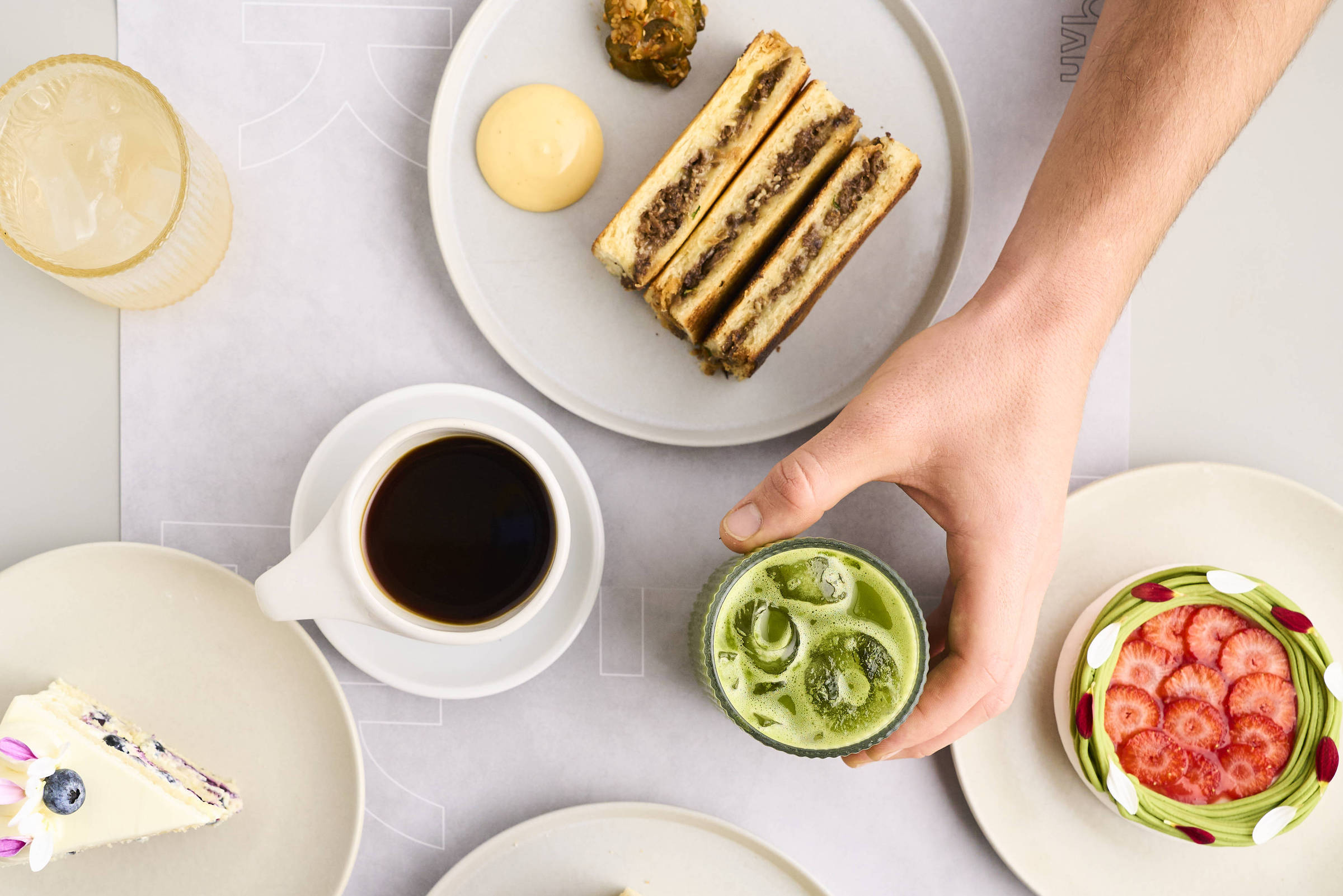 A imagem mostra uma mesa com vários pratos e bebidas. Há um prato com sanduíches, um copo com uma bebida verde, uma xícara de café, um prato com um doce e um prato com uma sobremesa colorida. As mãos de duas pessoas estão visíveis, uma segurando um sanduíche e a outra segurando o copo. O fundo é claro e a mesa é organizada.
