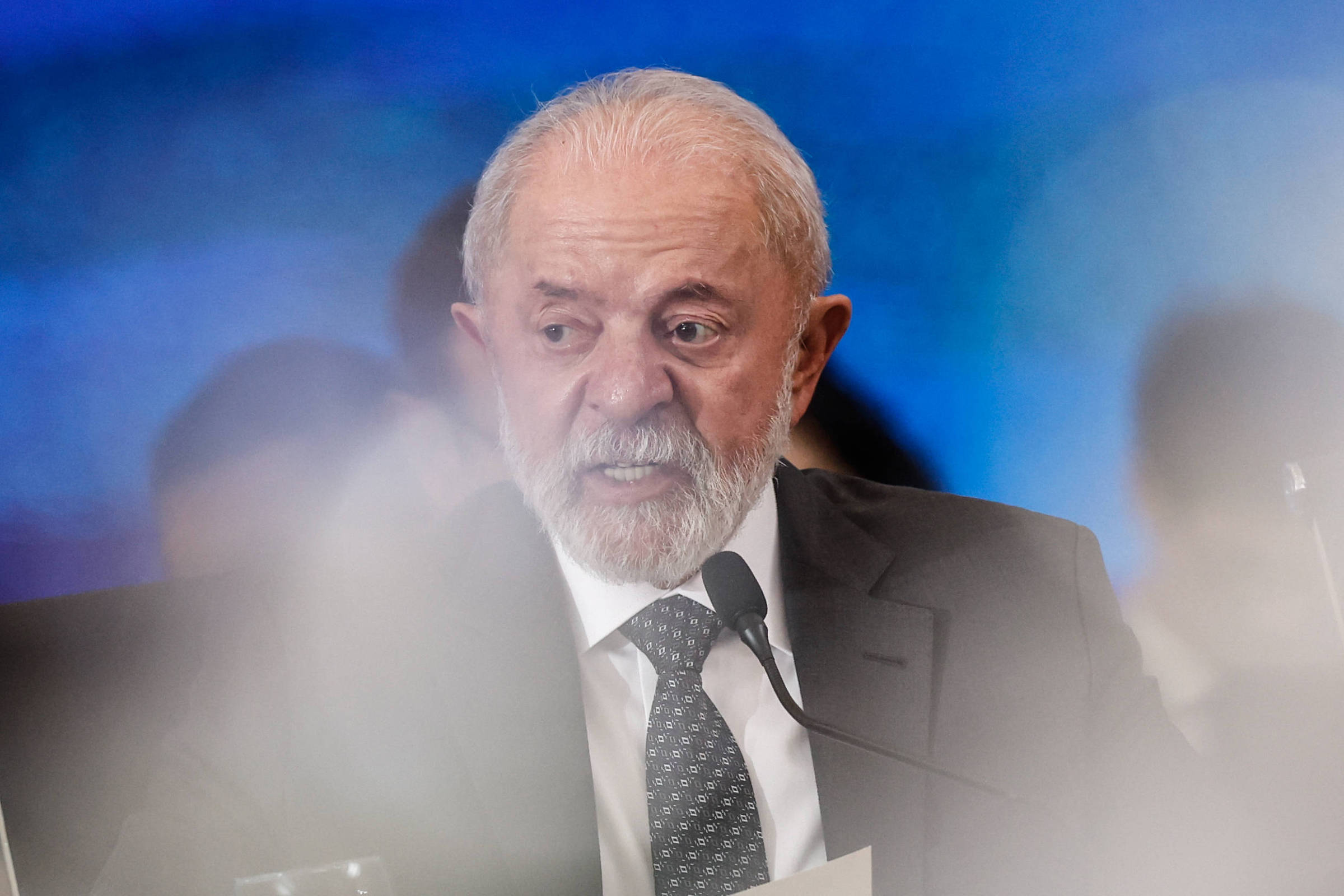 Um homem de cabelos grisalhos e barba, vestido com um terno escuro e gravata, está falando em um microfone. Ele parece estar em um evento formal, com um fundo desfocado que sugere uma plateia ou outros participantes. A expressão facial do homem é séria e concentrada, e ele segura um papel em uma das mãos.