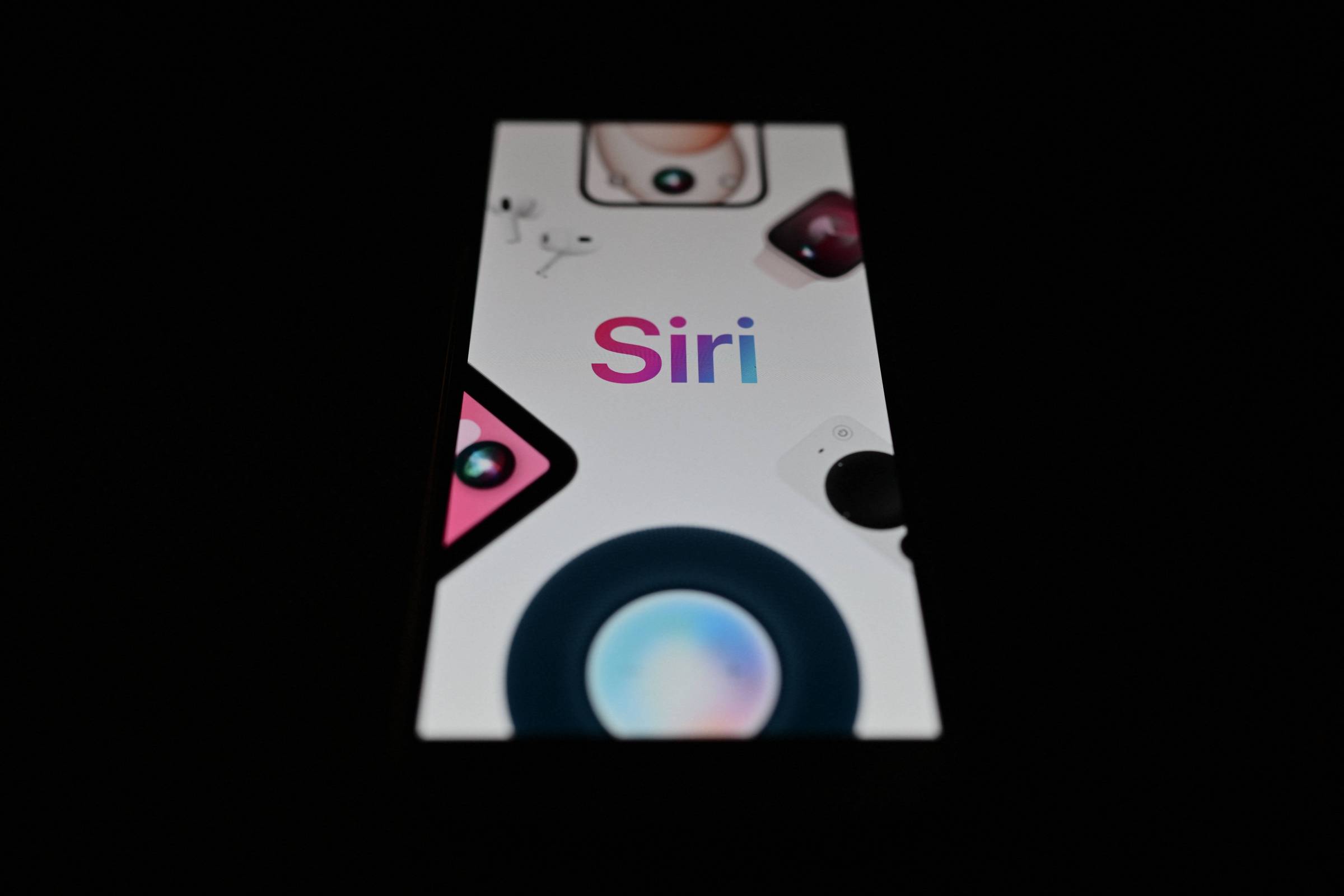A imagem mostra uma tela de smartphone com o logotipo do Siri em destaque. O fundo é claro, com ícones de dispositivos e elementos gráficos relacionados ao Siri, como câmeras e alto-falantes. A tela está iluminada em um ambiente escuro, destacando o texto colorido do logotipo.