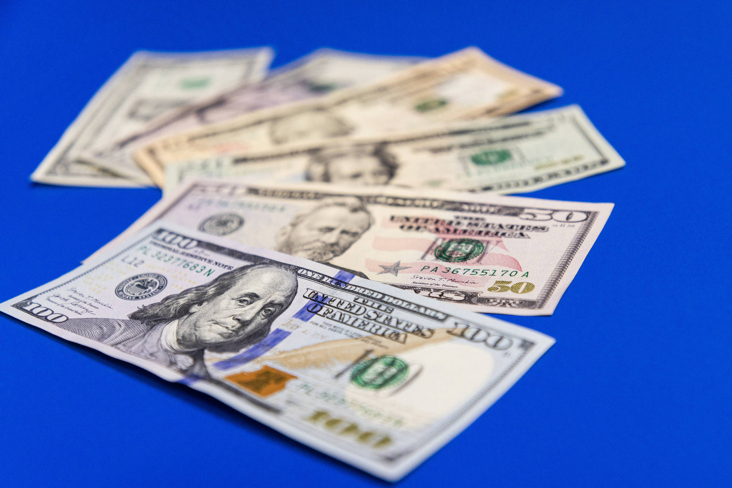 A imagem mostra várias notas de dólar dispostas sobre um fundo azul. As notas incluem diferentes denominações, com destaque para a nota de 100 dólares, que está em primeiro plano. As notas estão levemente sobrepostas, criando uma composição visual interessante.