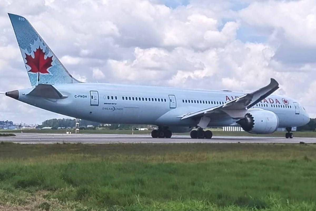 Um avião da Air Canada, modelo Boeing 787, está taxiando na pista de um aeroporto. O avião é de cor azul claro com detalhes em vermelho, incluindo uma folha de bordo no tail. O céu está parcialmente nublado, e há uma área verde ao primeiro plano.