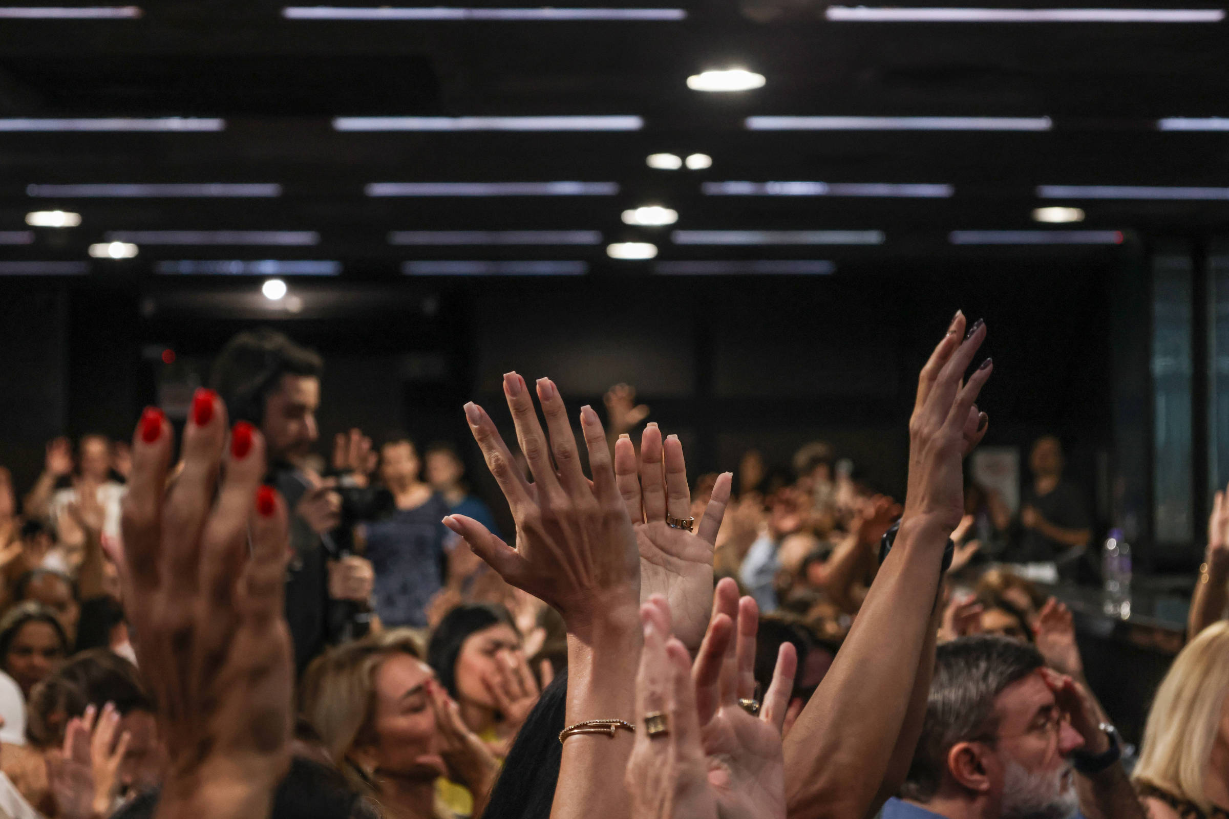 A imagem mostra uma multidão em um ambiente interno, com várias mãos levantadas. As pessoas parecem estar participando de um evento ou reunião, com expressões de entusiasmo. O fundo é escuro, com luzes de teto visíveis, e há uma variedade de tons de pele e estilos de unhas entre as mãos levantadas.