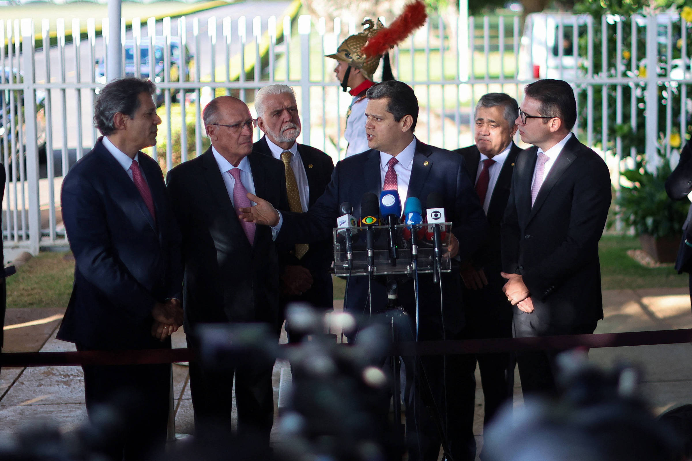 A imagem mostra um grupo de homens em um evento, possivelmente uma coletiva de imprensa. No centro, um homem está em pé em frente a um microfone, cercado por outros homens que parecem estar ouvindo atentamente. Há câmeras e jornalistas ao redor, indicando que é um momento de cobertura da mídia. Ao fundo, pode-se ver uma área verde e uma estrutura que parece ser um edifício.