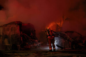A imagem mostra um bombeiro combatendo um incêndio em um local onde há veículos queimados. O ambiente está envolto em fumaça densa e há uma iluminação avermelhada, possivelmente das chamas. O bombeiro está usando equipamento de proteção e segurando uma mangueira, enquanto tenta controlar o fogo.