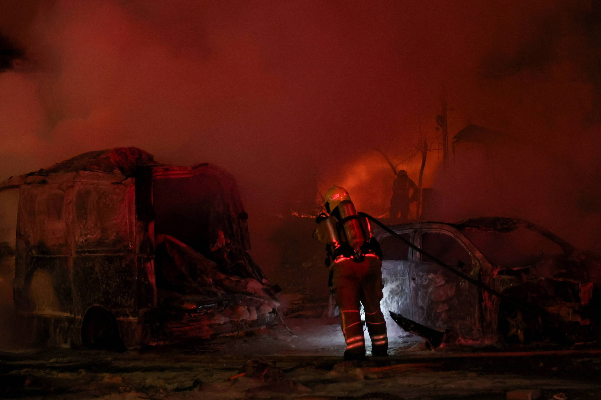 A imagem mostra um bombeiro combatendo um incêndio em um local onde há veículos queimados. O ambiente está envolto em fumaça densa e há uma iluminação avermelhada, possivelmente das chamas. O bombeiro está usando equipamento de proteção e segurando uma mangueira, enquanto tenta controlar o fogo.